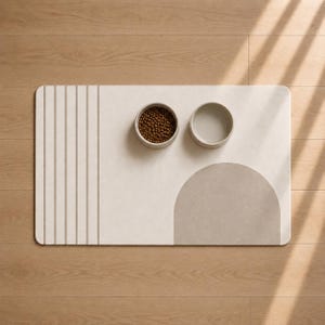 May include: A rectangular pet feeding mat in off-white featuring a series of vertical lines on the left and a large taupe semi-circle on the right. Two round bowls are placed on the mat, one filled with brown dry pet food and one empty.