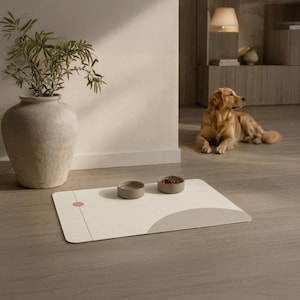 Puede incluir: Una alfombrilla rectangular para mascotas en color blanco roto con un diseño gráfico minimalista que incluye una línea vertical fina, un pequeño círculo rosa y un gran semicírculo beige. Dos cuencos redondos y claros están sobre la alfombrilla.