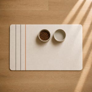 以下が含まれることがあります： 角が丸い長方形のオフホワイトのペット用フードマット。左側にグレーとオレンジの縦縞が3本入っています。マットの上には円形のボウルが2つ置かれており、片方には茶色のドライフードが入っており、もう片方は空です。