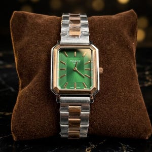 Pode incluir: Um relógio retangular com mostrador verde e pulseira de metal prateado e dourado. O relógio tem uma moldura dourada e é exibido em uma almofada de veludo marrom. O mostrador do relógio tem a marca "Tissot" impressa.