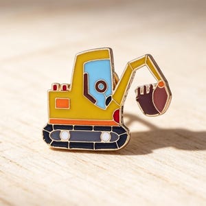 Könnte beinhalten: Ein gelber Bagger-Anstecker mit blauem Führerhaus und braunem Eimer. Der Anstecker hat schwarze, orangefarbene und rote Akzente. Der Bagger ist ein Accessoire zum Thema Bau.