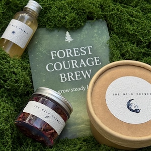 May include: A selection of products from "The Wild Brewer" displayed on green moss. The image features a small bottle, a jar with a silver lid, and a round container with a label. A card with the text "FOREST COURAGE BREW" is also visible.