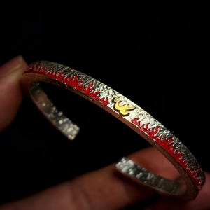 Puede incluir: Brazalete de plata con diseño de llamas rojas y amarillas. El brazalete tiene una superficie texturizada y un relleno de esmalte rojo brillante. Un emblema amarillo es visible en el brazalete.