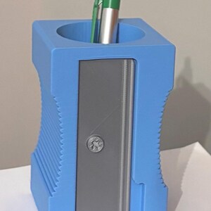 May include: A blue pencil holder shaped like a large pencil sharpener with a grey front panel and a single silver screw. A silver pen with a green clip is inserted into the circular opening at the top of the blue, textured, rectangular container.