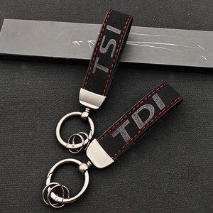May include: Two black keychains with silver metal hardware. The keychains have the text "TSI" and "TDI" printed in gray. Red stitching accents the edges of the black straps. The keychains are on a dark surface.