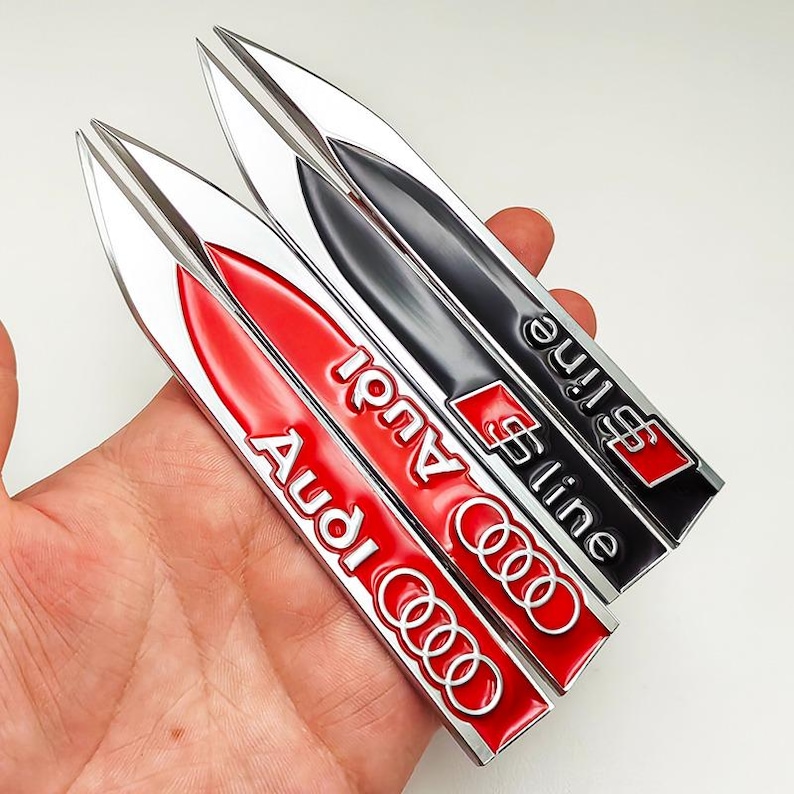 May include: Three car emblem badges. One red badge with the word "Audi" in white, and the Audi logo. One black badge with the words "S line" in silver and the S line logo. The badges are held in a hand.