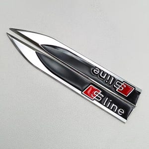 May include: Two silver and black S line car emblems. The emblems have a black background with the text "S line" in white and a red square with the "S" logo. The emblems are angled and have a pointed shape.