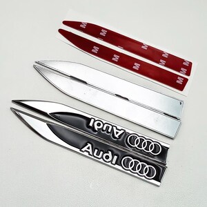 May include: A set of silver and black Audi car emblems. The black emblems have the Audi logo and text in white. Two silver emblems and two red adhesive strips are also visible.