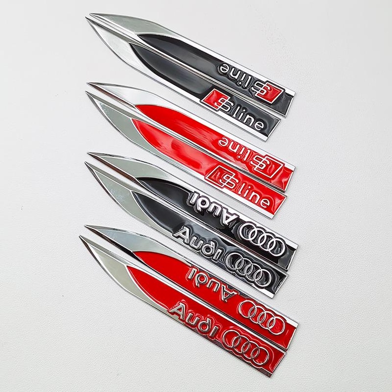May include: A collection of five car emblems in a sleek, elongated design. The emblems feature a silver and black or red color scheme, with the "S line" and "Audi" logos prominently displayed. These automotive accessories are designed for car enthusiasts.