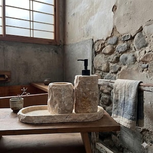 Puede incluir: Un juego de baño de piedra beige que incluye un dispensador de jabón con bomba negra, un vaso y una bandeja. El juego tiene una superficie rugosa y texturizada. Una pequeña toalla cuelga de una barra de madera.