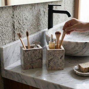 Peut inclure: Deux récipients carrés en pierre sur un comptoir en marbre contiennent des brosses à dents et des pinceaux de maquillage. Une bouteille de sérum pour le visage et un savon sont également visibles. Le lavabo et le robinet de la salle de bain sont en arrière-plan.
