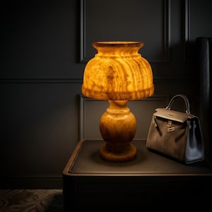 May include: An illuminated amber-toned table lamp with a rounded base and a flared shade. It's placed on a dark brown table, beside a brown leather handbag with gold details. The backdrop is a dark, textured wall.