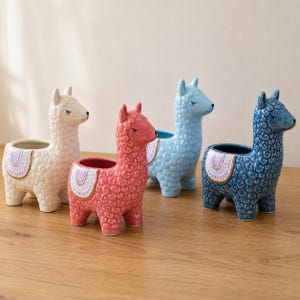 Puede incluir: Cuatro maceteros de cerámica con forma de llama en blanco, rosa, azul claro y azul oscuro. Cada macetero presenta un pelaje rizado texturizado, ojos cerrados y una manta de silla decorativa con patrones morados y blancos en el lomo.