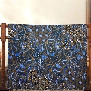 Op de afbeelding: Een patroontextiel in donkerblauw en goud wordt op een houten rek getoond. De stof heeft een complex ontwerp met bloem- en abstracte vormen. Een bruine vaas met gedroogde bloemen en andere decoratieve items staan op de achtergrond.