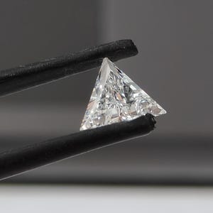 May include: A clear, colorless, triangular-cut gemstone with multiple facets that reflect light. The stone is held by black tweezers, showcasing its sharp, geometric shape and transparent, crystalline structure against a neutral background.