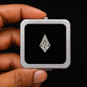 Puede incluir: Una piedra preciosa transparente con facetas en forma de cometa, presentada dentro de un estuche metálico cuadrado de color plateado con un interior de terciopelo negro. La piedra tiene facetas geométricas simétricas.