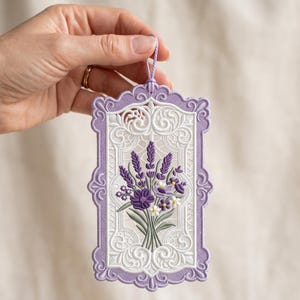 Puede incluir: Un adorno colgante bordado rectangular con un diseño de ramo de flores en lavanda y blanco. La pieza tiene un borde festoneado de color lavanda con intrincados patrones de filigrana blancos y un cordón de color lavanda a juego.