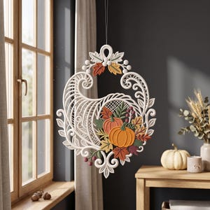 Può includere: Un ornamento bianco a forma di cornucopia dal design intricato simile al pizzo, appeso a un filo sottile. Presenta un motivo intrecciato con bordi in filigrana e un gruppo centrale di elementi autunnali ricamati, tra cui zucche arancioni, foglie verdi e rosse e piccole bacche.