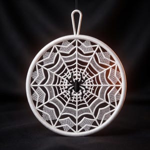 May include: A circular white ornament featuring an intricate spiderweb pattern with a solid black spider positioned in the center. The design includes a top loop for hanging, and the entire structure is rendered in a clean, matte finish.