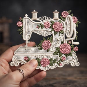 May include: An embroidered patch shaped like a vintage sewing machine in cream thread with a cross-stitch pattern. The design is decorated with detailed pink roses and green leaves, featuring two thread spools on top and a hand crank on the side.
