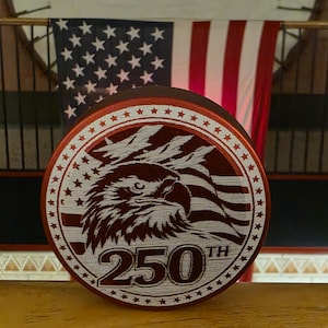 Puede incluir: Un objeto conmemorativo redondo, rojo y blanco, con un águila, la bandera estadounidense y el texto "250th". El diseño está grabado. Una bandera estadounidense está en el fondo.