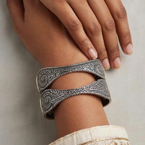 May include: Two silver-toned cuff bracelets featuring intricate, embossed scrollwork and filigree patterns. The bracelets have a curved, open-work design with a darkened, oxidized finish that highlights the detailed metal engravings across the surface.