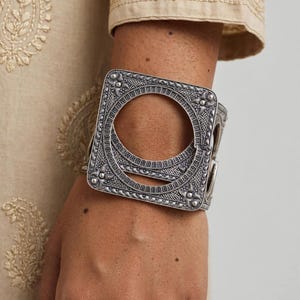 May include: A wide, silver-toned cuff bracelet featuring an intricate, oxidised metal finish with a prominent square frame design. The piece showcases detailed filigree patterns, small spherical accents, and a large circular cutout at the centre.