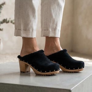 May include: Black suede clogs featuring a plush, dark-colored interior lining, a rounded toe, and a thick wooden heel. The base of the shoe is secured with a row of small, evenly spaced metal studs along the perimeter of the light-colored wooden sole.