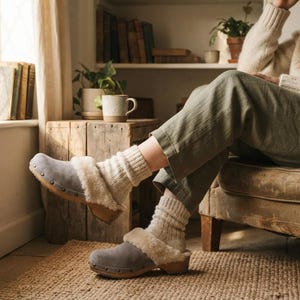 May include: Grey suede clogs featuring a light-colored faux fur lining and a wooden sole. The clogs are worn with thick, ribbed, cream-colored knit socks. The footwear has a rounded toe design and visible metal studs securing the upper to the base.