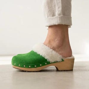 May include: A bright green suede clog featuring a thick white shearling lining around the collar. The shoe has a light-colored wooden sole and heel, accented with a row of small silver-toned metal studs along the base of the upper material.