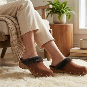 May include: A pair of brown suede clogs featuring a dark brown faux fur lining, a wooden sole, and a row of silver-toned metal studs along the base of the upper. The slip-on footwear has a rounded toe and a low heel, providing a classic profile.