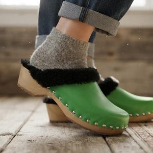 May include: A pair of bright green leather clogs featuring a light-colored wooden sole, a black faux fur lining along the collar, and a row of silver-toned metal studs securing the leather upper to the wooden base of the footwear.