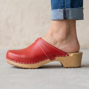 Puede incluir: Un zueco de cuero rojo con acabado liso y suela de madera clara. El zapato presenta una puntera redondeada, una tira ancha sobre el empeine y costuras blancas visibles a lo largo de los bordes de la parte superior de cuero, sobre un tacón bajo.