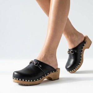 Pode incluir: Tamancos de couro preto com um detalhe de bridão em metal prateado no peito do pé. Os sapatos têm uma sola e salto de madeira clara, acentuados com uma fila de pequenos rebites de metal prateado ao longo da base do cabedal de couro.