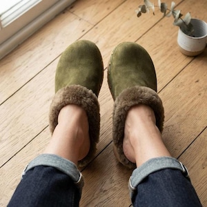 May include: A pair of olive green suede clogs featuring a rounded toe and a thick, plush brown faux fur lining that extends around the collar. The clogs have a sturdy construction with visible small metal studs along the side of the sole.