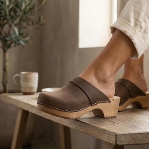 May include: A pair of brown leather clogs featuring a wide strap across the top, a rounded toe, and a light-colored wooden sole with a low heel. The leather upper is secured to the wooden base with a row of small metal studs along the side.