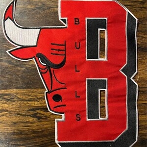 Puede incluir: Parche rojo y negro con el logotipo de los Chicago Bulls y el número 3. El logotipo incluye una cabeza de toro roja con detalles en blanco. La palabra "BULLS" está bordada en blanco sobre el fondo rojo.
