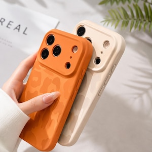 May include: Two phone cases, one orange and one cream, are displayed. The orange case has a leopard print design and is held in a hand. The cream case also has a leopard print design. Both cases have camera cutouts.