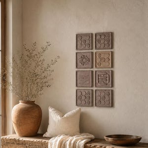 May include: A rustic wooden bench with a cream-coloured throw blanket and a pillow, a large terracotta vase with dried branches, a dark wooden bowl, a woven basket, and eight square wall tiles featuring embossed geometric and floral patterns.