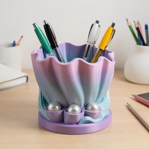 Puede incluir: Un portalápices con forma de concha marina, impreso en 3D con un degradado de rosa, morado y azul. El soporte tiene tres detalles en forma de perla y contiene varios bolígrafos de diferentes colores.