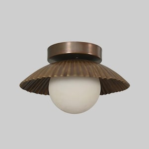 May include: A flush mount ceiling light fixture featuring a circular bronze base and a wide, fluted, scalloped bronze shade. A single, spherical, opaque white glass globe bulb is centered beneath the metal shade, providing a balanced and symmetrical appearance for this lighting piece.
