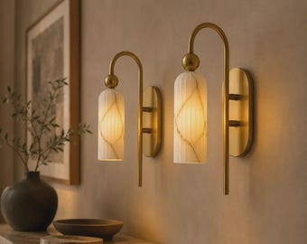 May include: Two wall sconces featuring a curved brass arm and a cylindrical, vertically ribbed white glass shade with gold veining. Each light is mounted on an oval brass backplate and emits a warm, soft glow from the illuminated glass fixture.