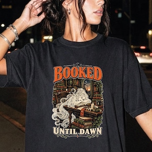 May include: A black short-sleeve t-shirt featuring a graphic print of a ghost reading a book at a desk in a library. The text "BOOKED" appears in orange above the illustration, and "UNTIL DAWN" is printed in white below the graphic design.