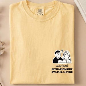 May include: A pale yellow short-sleeve t-shirt featuring a black and white graphic on the lower right side. The graphic depicts two figures with a question mark and the text: undefined SITUATIONS STATUS: MAYBE. The shirt has a crew neckline.