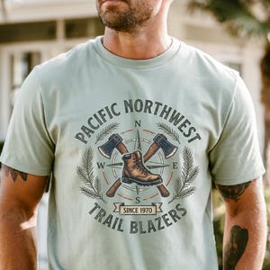 May include: A light sage green short-sleeve t-shirt featuring a graphic print of a brown hiking boot centered over two crossed axes. The design includes a compass rose, pine branches, and the text Pacific Northwest Trail Blazers Since 1970.