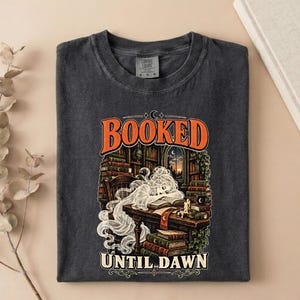 May include: A charcoal gray crew neck t-shirt featuring a graphic print of a ghost sleeping at a desk in a library. The text reads BOOKED UNTIL DAWN. The illustration includes stacks of books, a candle, and a window showing a sunset scene.