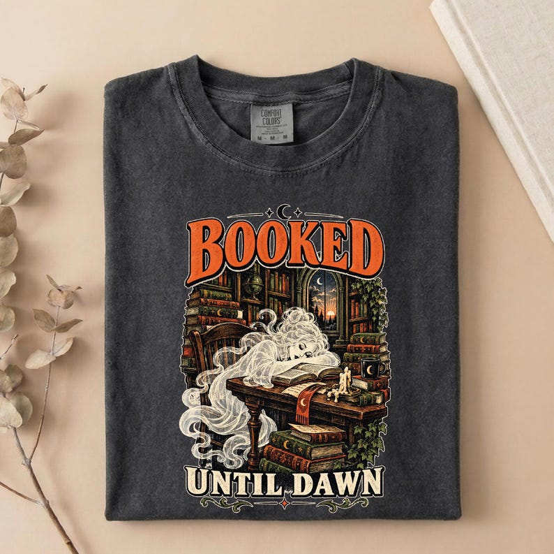 May include: A charcoal gray crew neck t-shirt featuring a graphic print of a ghost sleeping at a desk in a library. The text reads BOOKED UNTIL DAWN. The illustration includes stacks of books, a candle, and a window showing a sunset scene.
