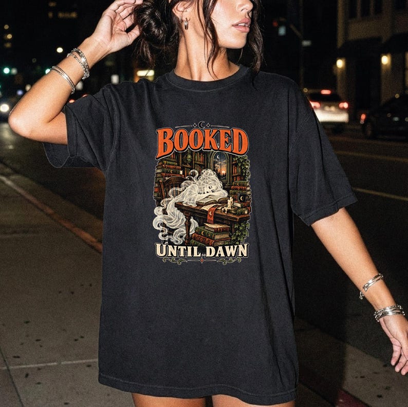May include: A black short-sleeve t-shirt featuring a graphic print of a ghost reading a book at a desk in a library. The text "BOOKED" appears in orange above the illustration, and "UNTIL DAWN" is printed in white below the graphic design.