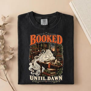 May include: A black crew neck t-shirt featuring a graphic print of a ghost reading a book at a desk in a library. The text "BOOKED" appears in orange above the illustration, and "UNTIL DAWN" is printed in white at the bottom of the design.