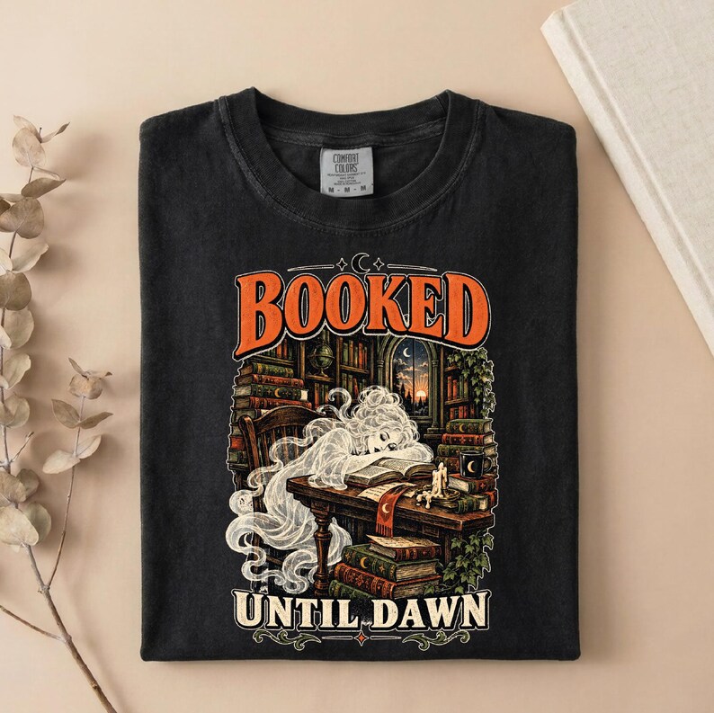 May include: A black crew neck t-shirt featuring a graphic print of a ghost reading a book at a desk in a library. The text "BOOKED" appears in orange above the illustration, and "UNTIL DAWN" is printed in white at the bottom of the design.